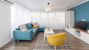 Una sala de estar moderna con un sofá azul, un sillón amarillo, una mesita y un televisor sobre una estantería de madera.