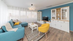Una sala moderna con un sofá azul, un sillón amarillo, una mesa de centro blanca y un televisor sobre un mueble de madera.