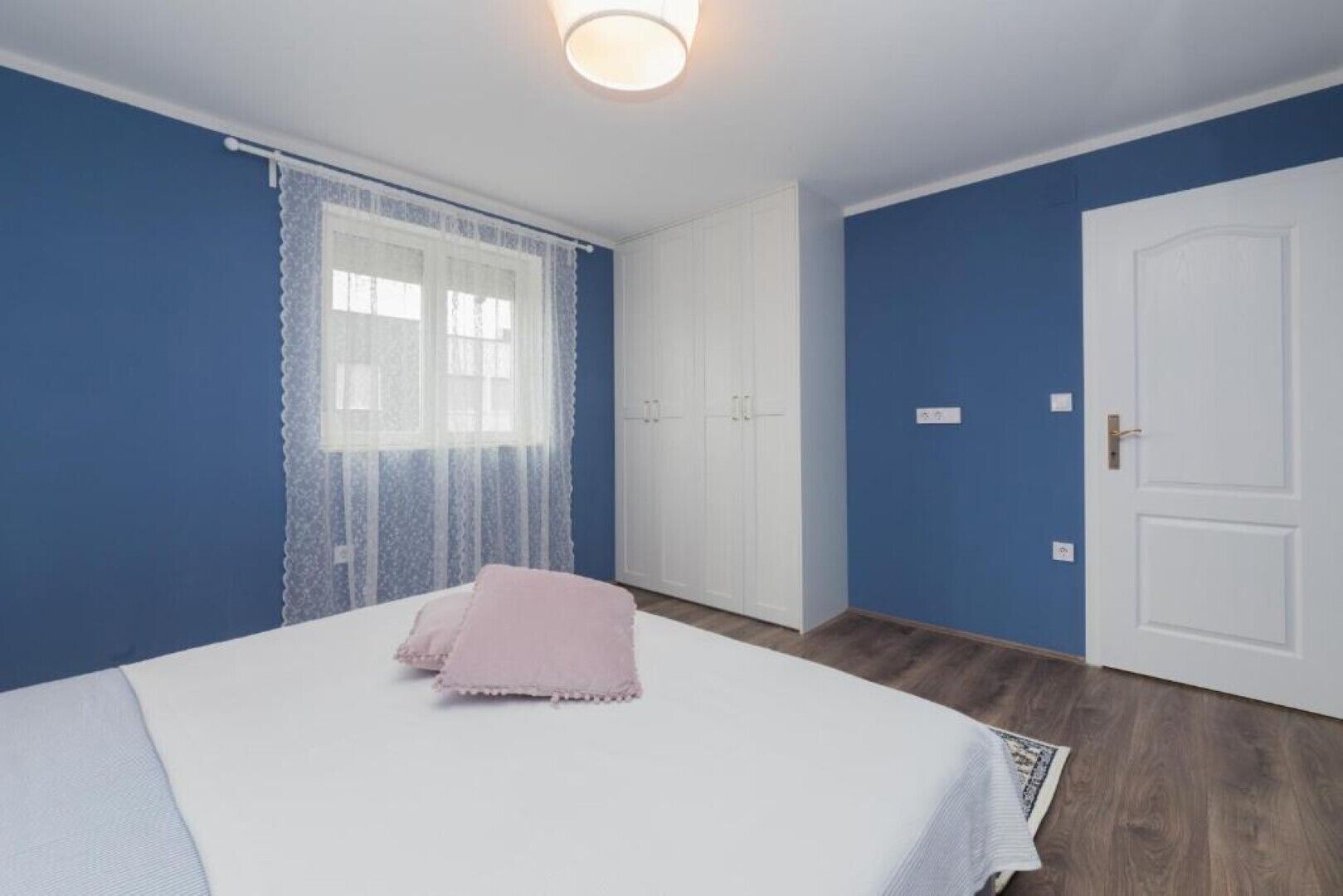 A bedroom with a bed, a window with lace curtains, a wardrobe, and a door.