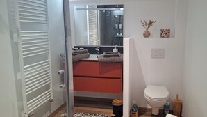 A modern bathroom with a glass shower enclosure, a red vanity, a white toilet, and a small wooden side table with toiletries. - B&B Baie du Mont Saint-Michel (Le Val-Saint-Père)