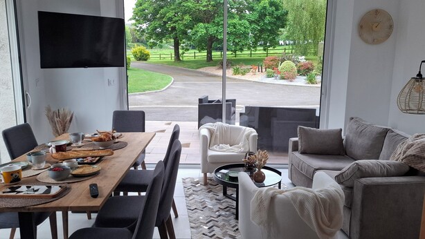 A modern living room with a dining area, large windows with a view of a green landscape, and contemporary furniture. - B&B Baie du Mont Saint-Michel (Le Val-Saint-Père)