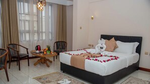 Quarto de hotel com cama, duas cadeiras, uma mesa redonda pequena com chaleira e fruta, e janela com cortinas.