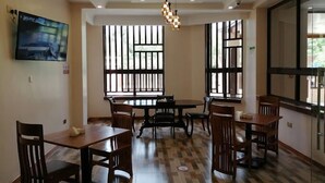 Uma sala de jantar com várias mesas e cadeiras, janelas amplas com persianas verticais, iluminação embutida no teto e uma televisão de tela plana na parede.