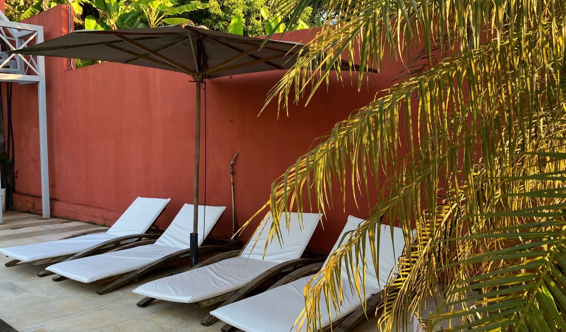 A row of sun loungers with white cushions, a metal umbrella, and a red wall.
