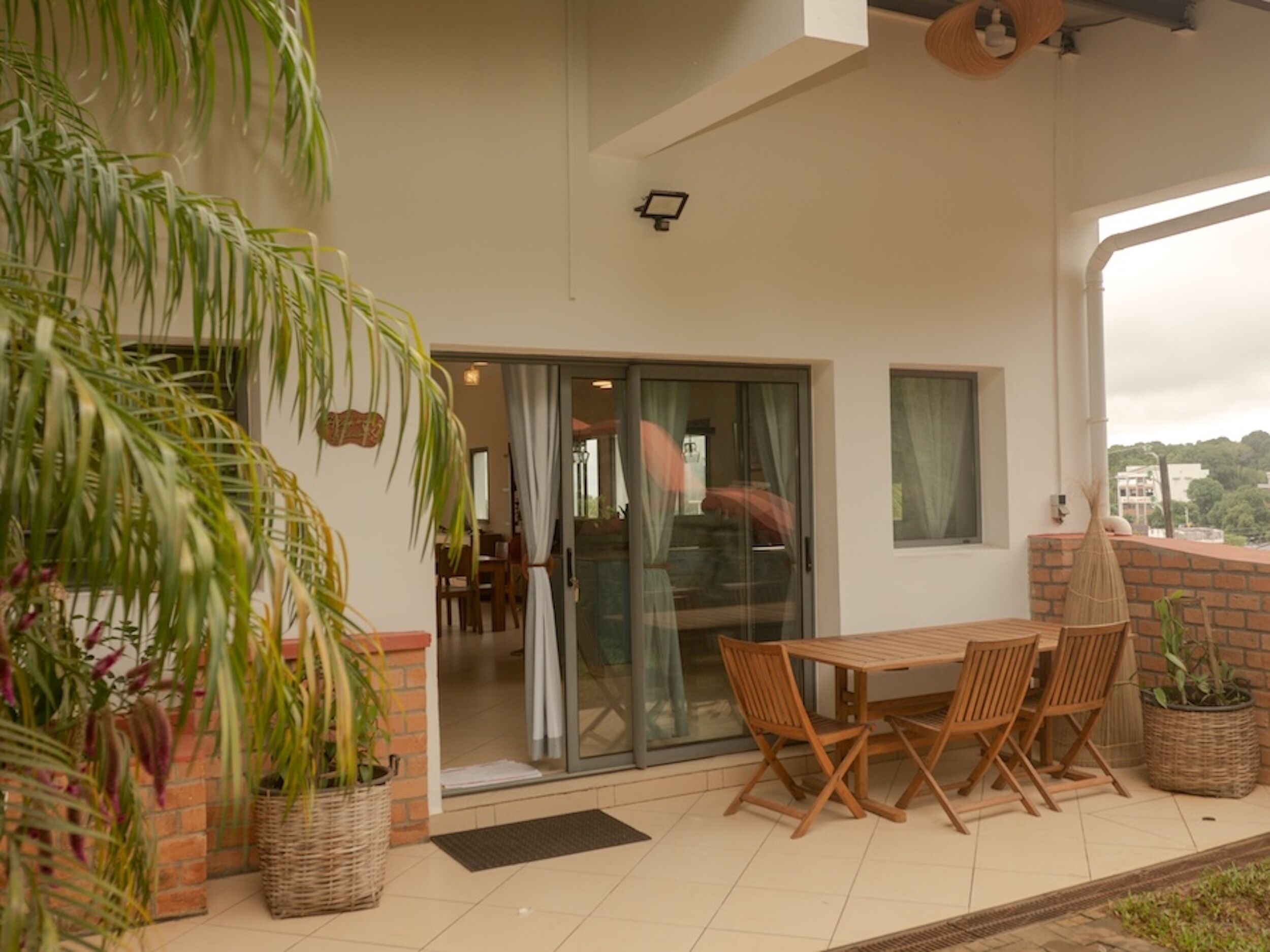 Un patio con una mesa y sillas de madera, plantas en macetas y un ventanal corredero que da a un espacio interior.