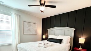 A neatly made bed with a white quilt, a ceiling fan, a bedside table with a lamp, and a window with curtains. - GIG ’EM LANE • 3BR • Grill & Fire Pit • Coffee Bar • 75" TV • Near Hwy 6 (College Station)