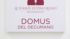 Een bordje met de tekst 'LE STANZE DI VINO ROSSO BED & BREAKFAST DOMUS DEL DECUMANO', met een icoontje van een wijnglas.