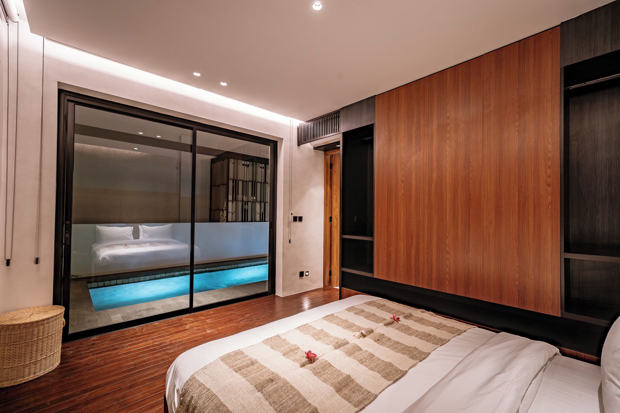 A modern bedroom with a large bed, a sliding glass door leading to a pool area, and a wooden panel wall.