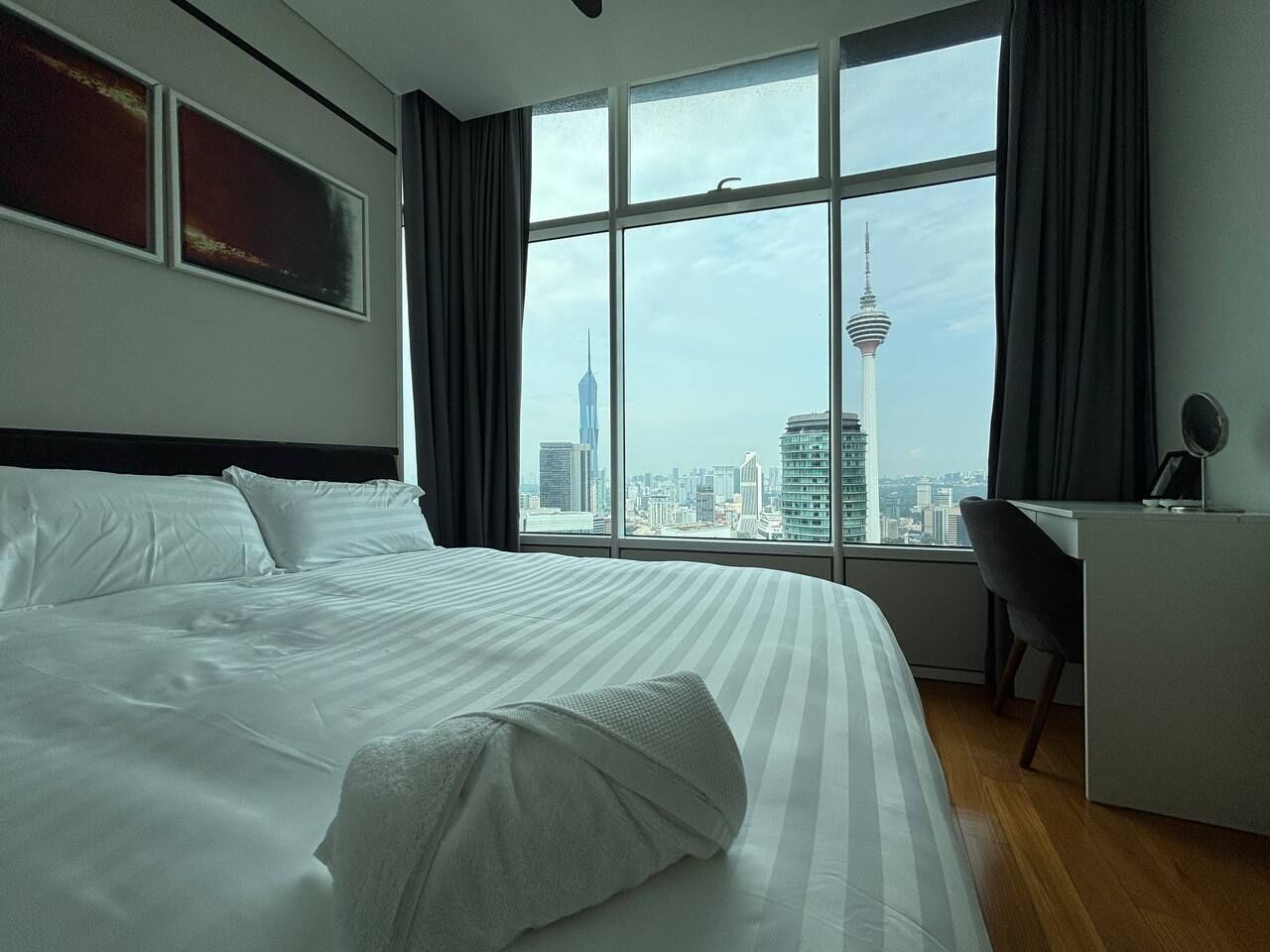 A hotel room with a large bed, a desk, and a view of a cityscape through large windows.
