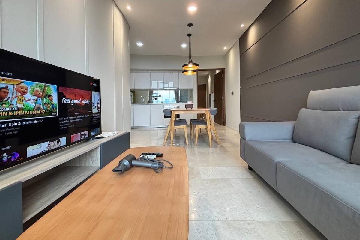 A modern living room with a flat-screen TV, a wooden coffee table, a grey sofa, and a dining area with a wooden table and chairs.