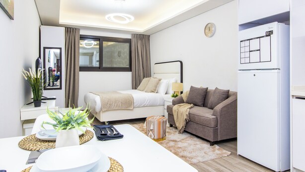A modern hotel room with a bed, a sofa, a dining table, and a refrigerator.