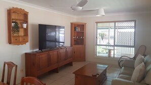 A living room with a TV, wooden cabinets, a ceiling fan, a sofa, and a window with blinds. - BANYA BLISS - quiet, cosy unit close to beach, cafes, bowls club and RSL (Bongaree)