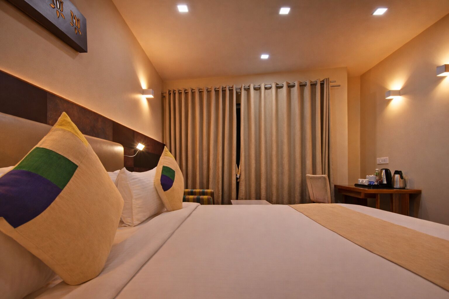 Quarto de hotel com uma cama grande, cabeceira, escrivaninha com cadeira e janela com cortinas.
