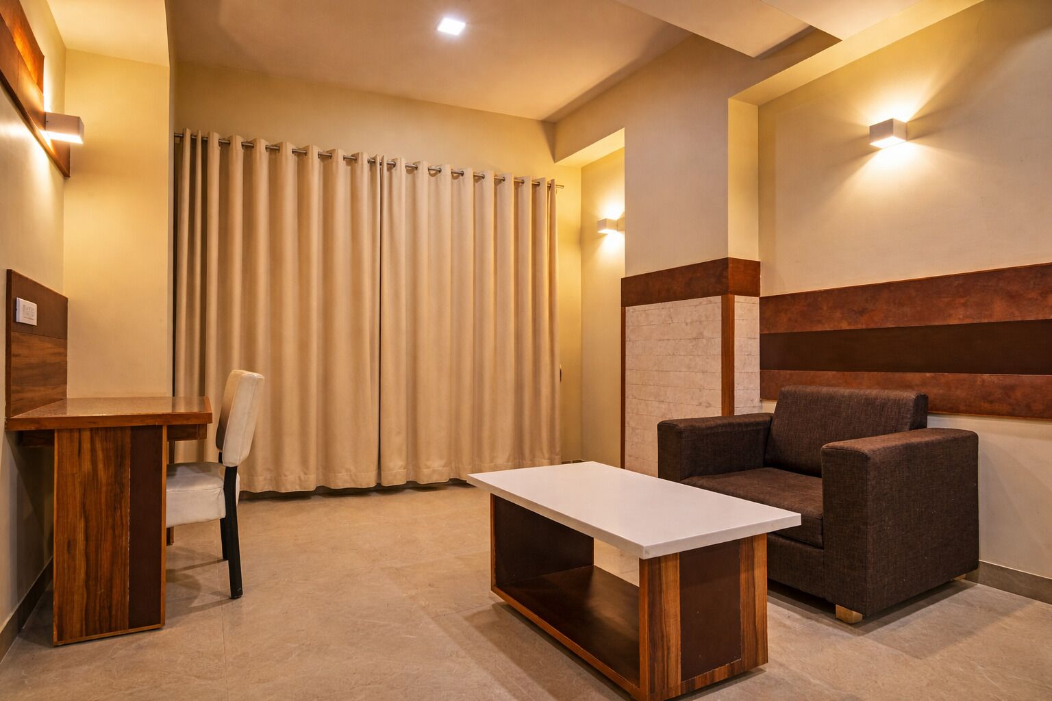 A hotel room with a wooden desk, a cushioned chair, a sofa, and a large window with curtains.