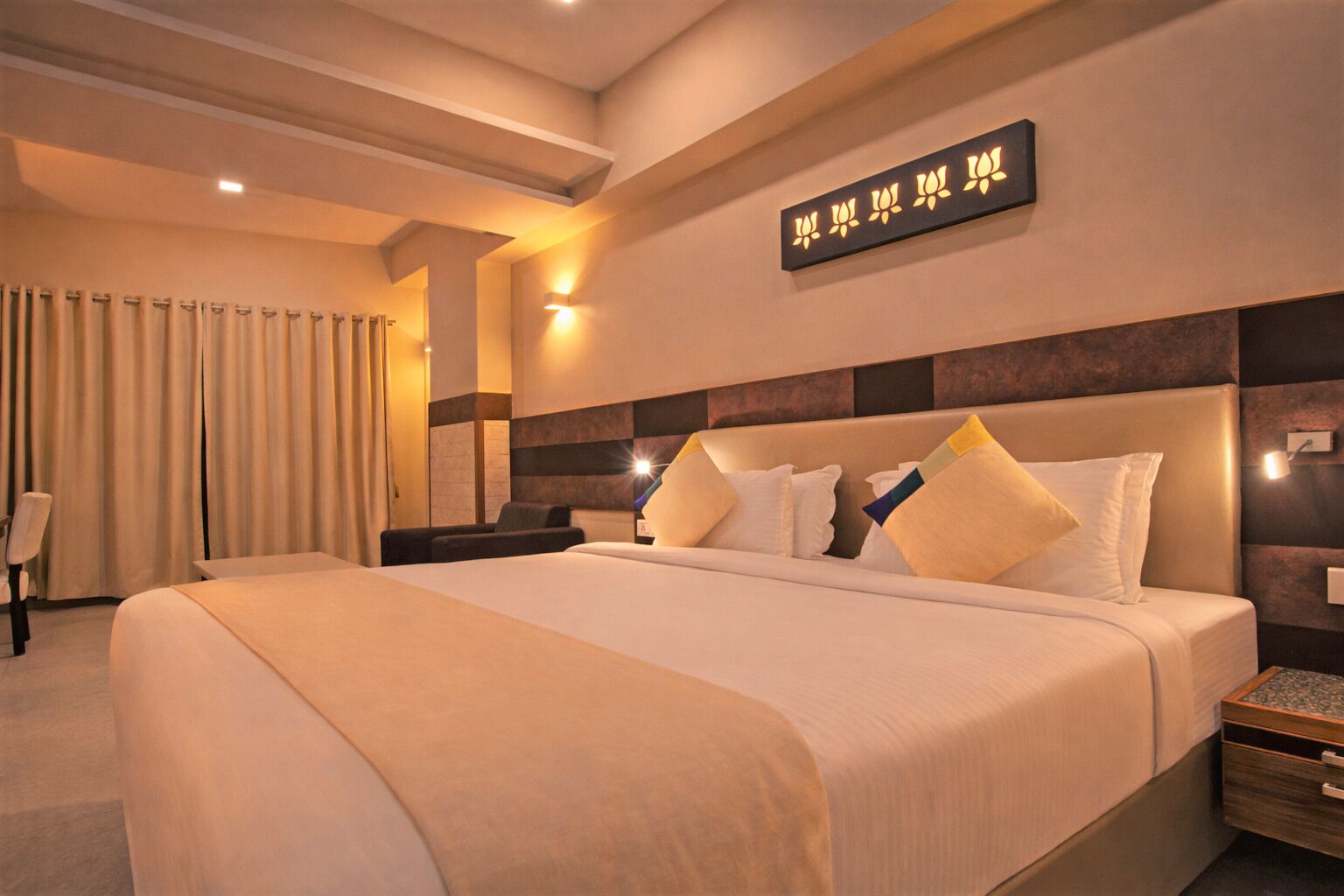 A large bed with multiple pillows, a bedside table, a wall-mounted light, and a decorative wall piece.