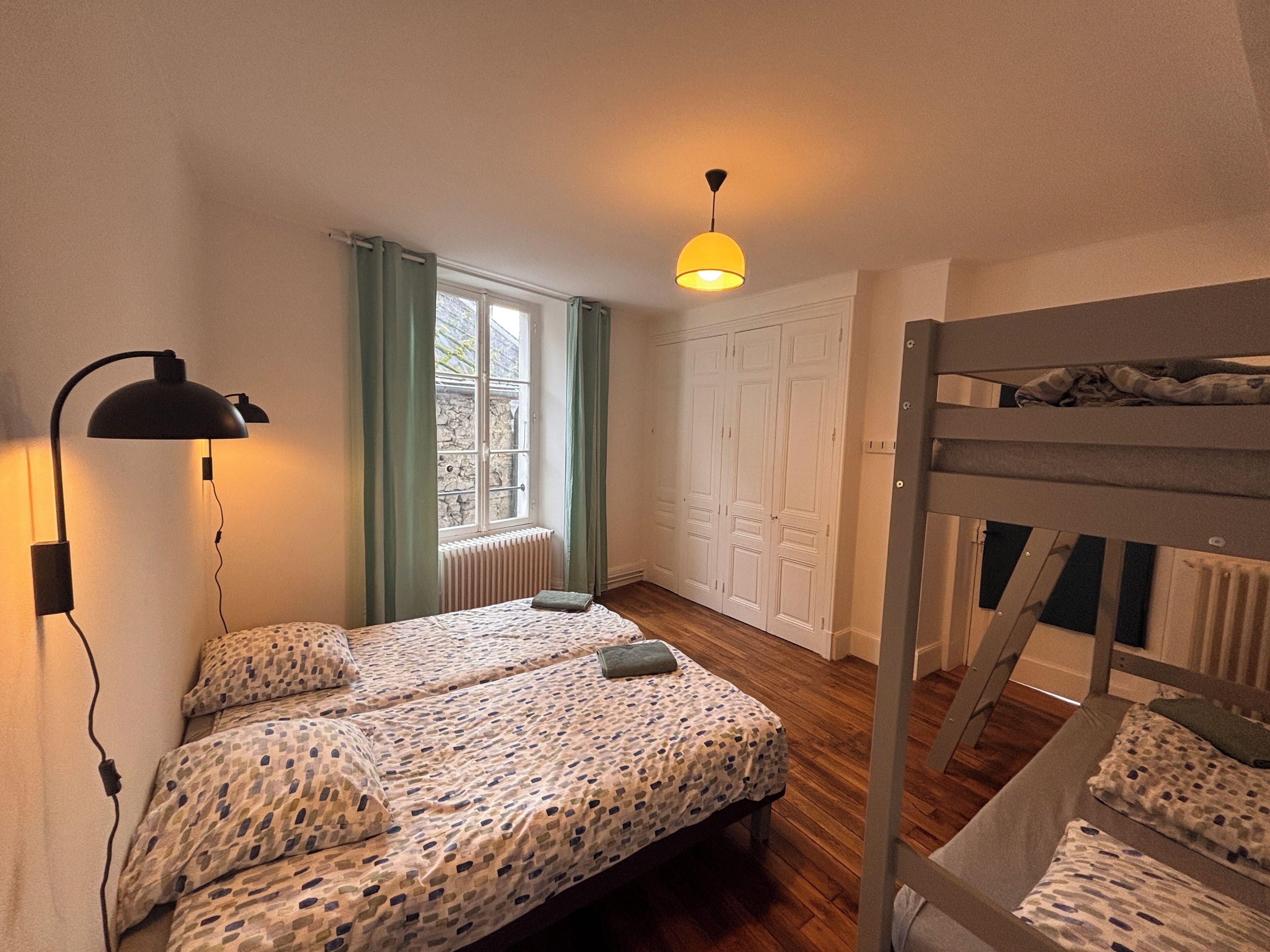 Ein kleines, gut beleuchtetes Schlafzimmer mit Etagenbett, Nachttischlampe, einem Fenster mit Vorhängen und einem Heizkörper.