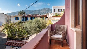 Un balcón con una silla de mimbre, con vista a una calle con edificios tradicionales y montañas al fondo.