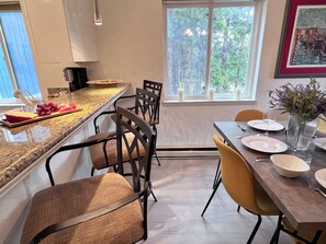 A modern kitchen with a marble countertop, a dining area with chairs, and a window with a view of trees.
