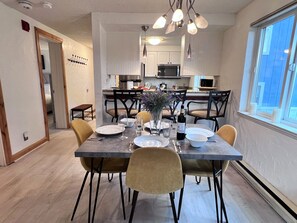 A dining area with a table set for four, chairs, a vase with flowers, and a kitchen in the background. - Mark IX Alpine Condo • Cozy Mtn Condo • 1 mile to Breck Main St + Slopes (Breckenridge)