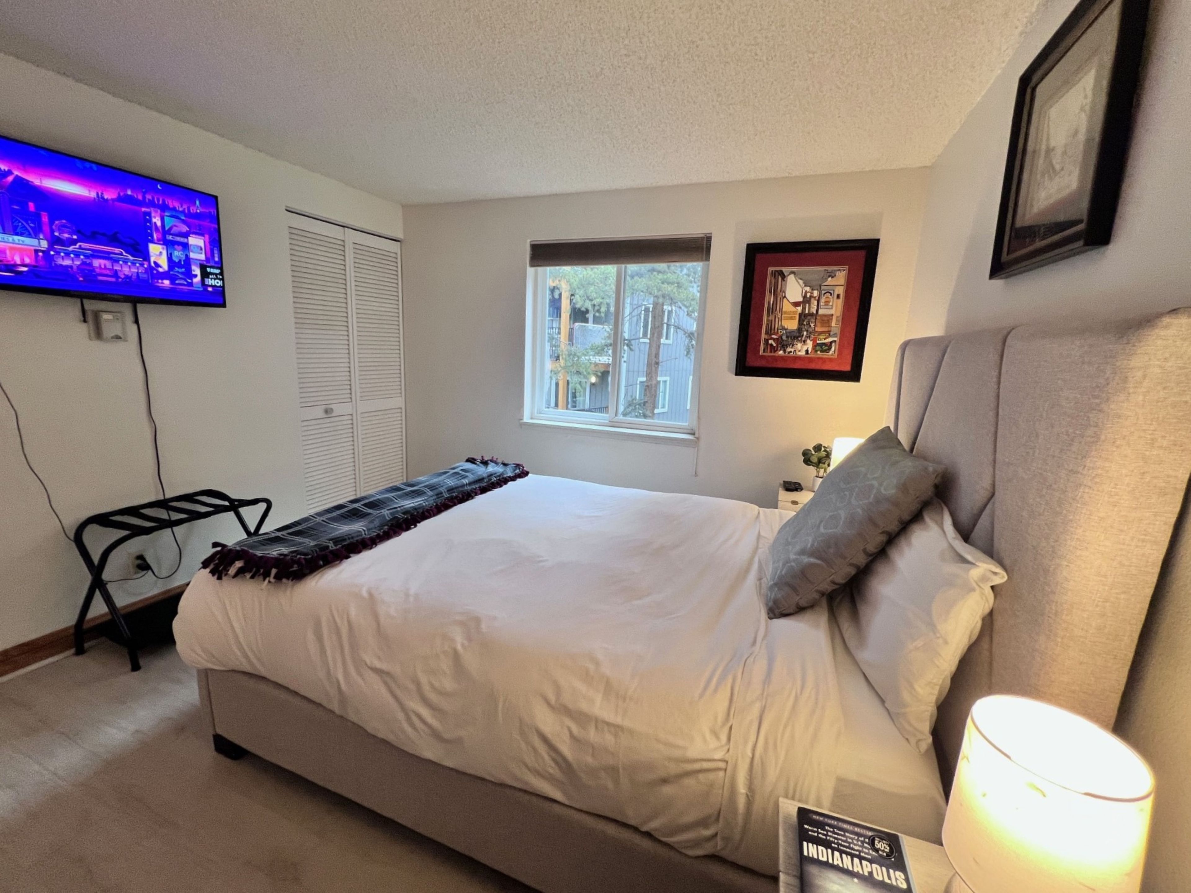 Un dormitorio con cama, televisor montado en la pared, ventana y un cuadro enmarcado.