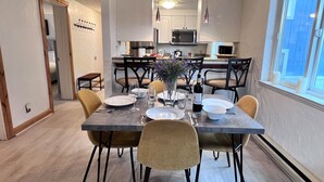 Un coin repas avec une table dressée pour quatre personnes, des chaises, un vase avec des fleurs et, au fond, une cuisine.