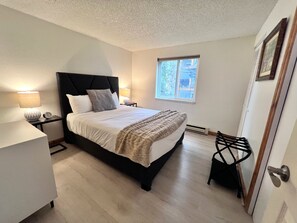A bedroom with a bed, bedside tables, a chair, a window, and a door. - Mark IX Alpine Condo • Cozy Mtn Condo • 1 mile to Breck Main St + Slopes (Breckenridge)