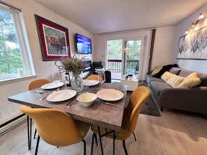 Un comedor con una mesa dispuesta para cuatro personas, un sofá, un televisor y un ventanal con vista a árboles.