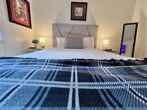 Una cama bien hecha con una colcha de cuadros, dos mesitas de noche con lámparas, un cuadro enmarcado en la pared y un espejo.