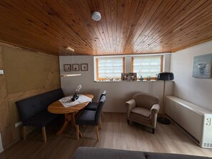 A cozy room with a wooden ceiling, a dining table with chairs, a sofa, a chair, a floor lamp, and a radiator.