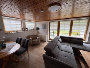 A modern living room with a wooden ceiling, a round table with chairs, a sofa, and large windows with blinds.