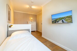 A bedroom with a bed, a dresser, a nightstand, a wall-mounted TV, and a door leading to another room. - Schaumkrönchen - Apartment Schaumkrönchen: Close to the beach with balcony. (Sylt)