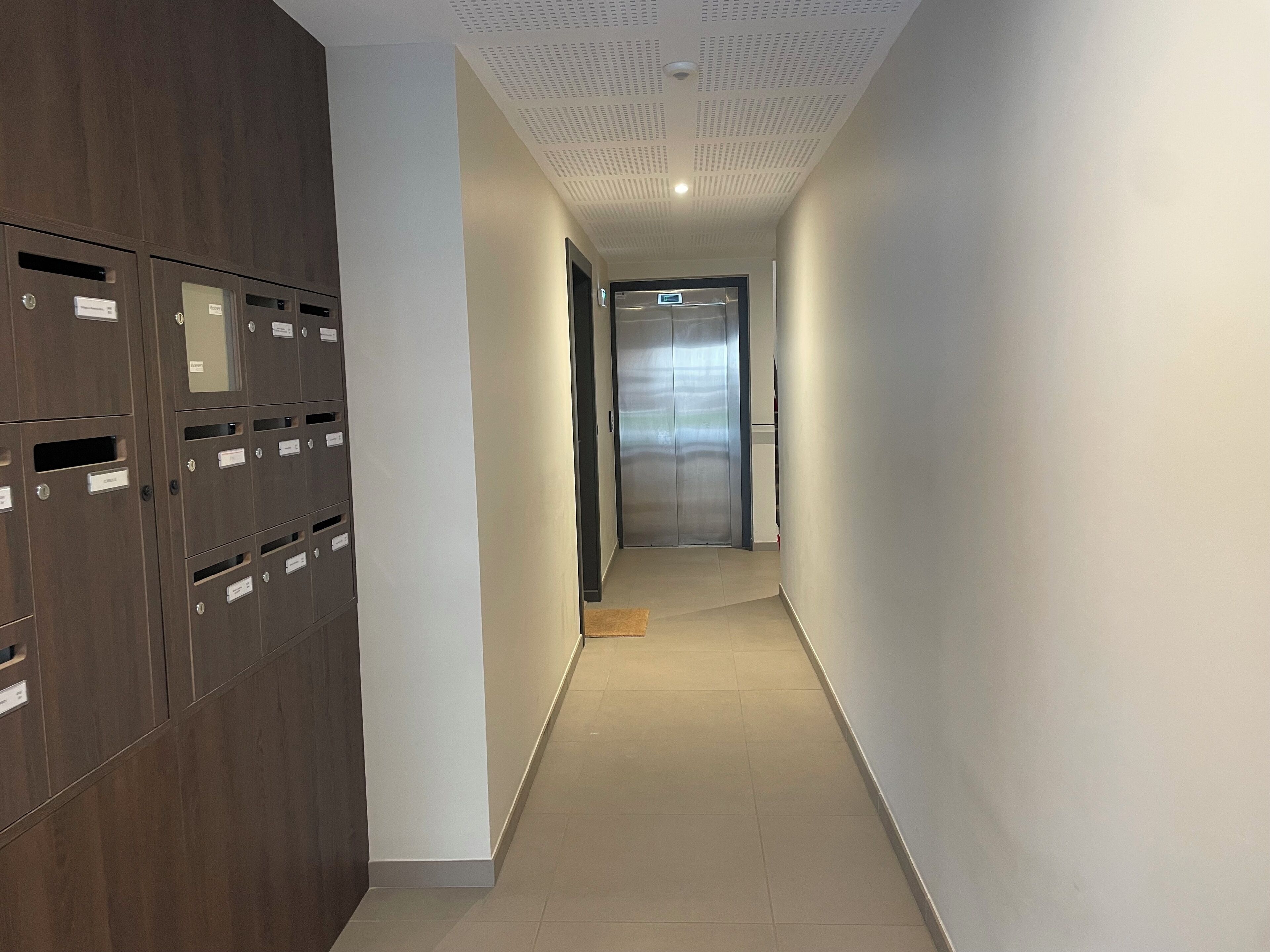 A hallway with lockers on the left, a door at the end, and a ceiling with a grid pattern.