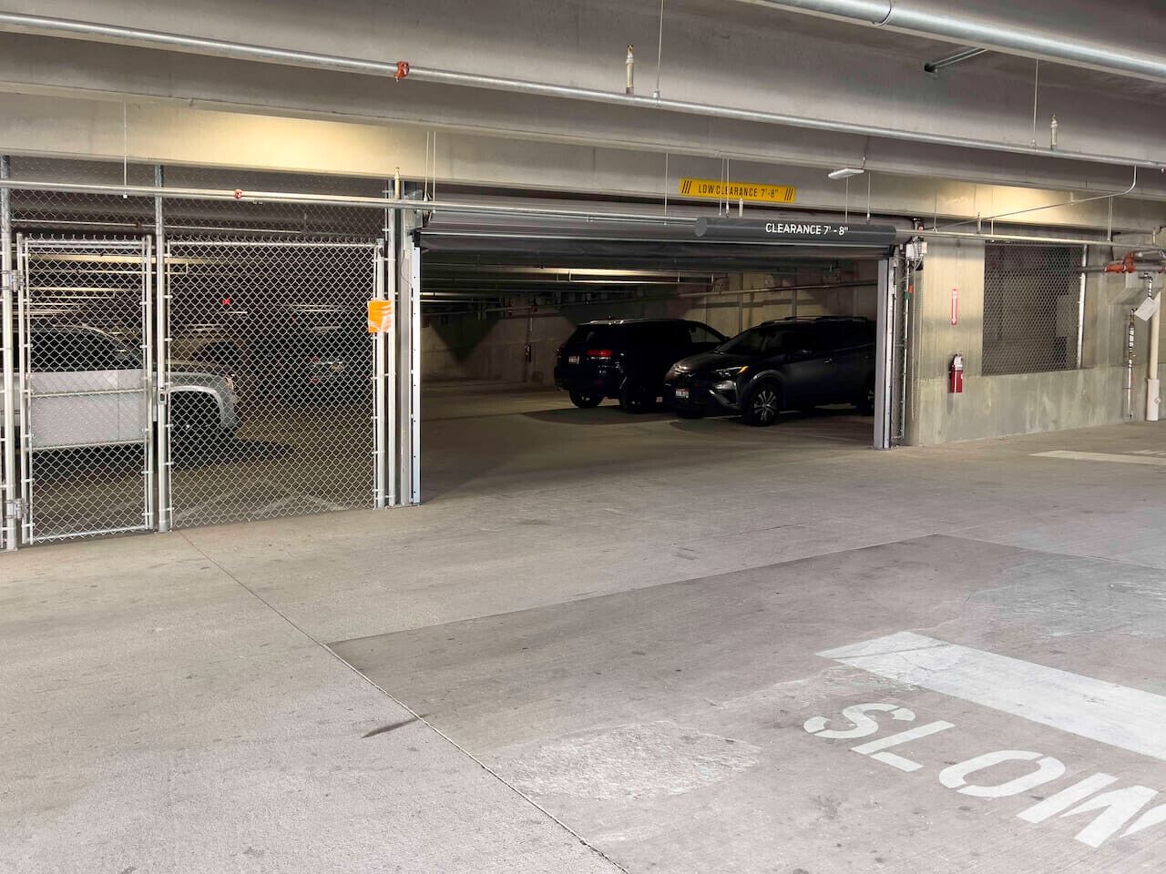 Un garaje de estacionamiento con puertas metálicas, una señalización de 'LENTO' en el suelo y un letrero de 'ALTURA 2.13-2.44 metros'.