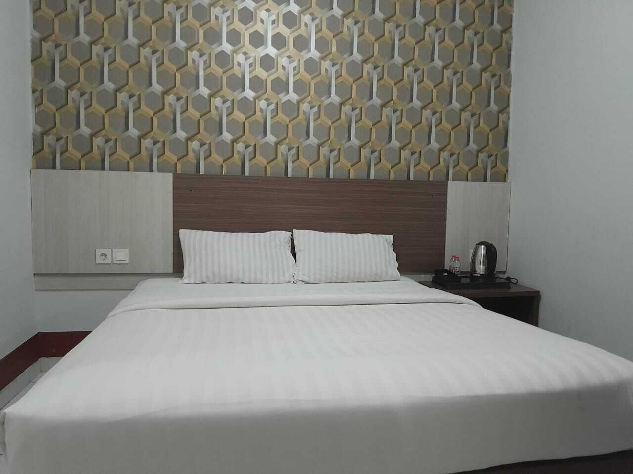 Habitación de hotel con una cama grande, cabecera de madera, panel decorativo en la pared, mesita de noche con lámpara y una pequeña mesa con hervidor.
