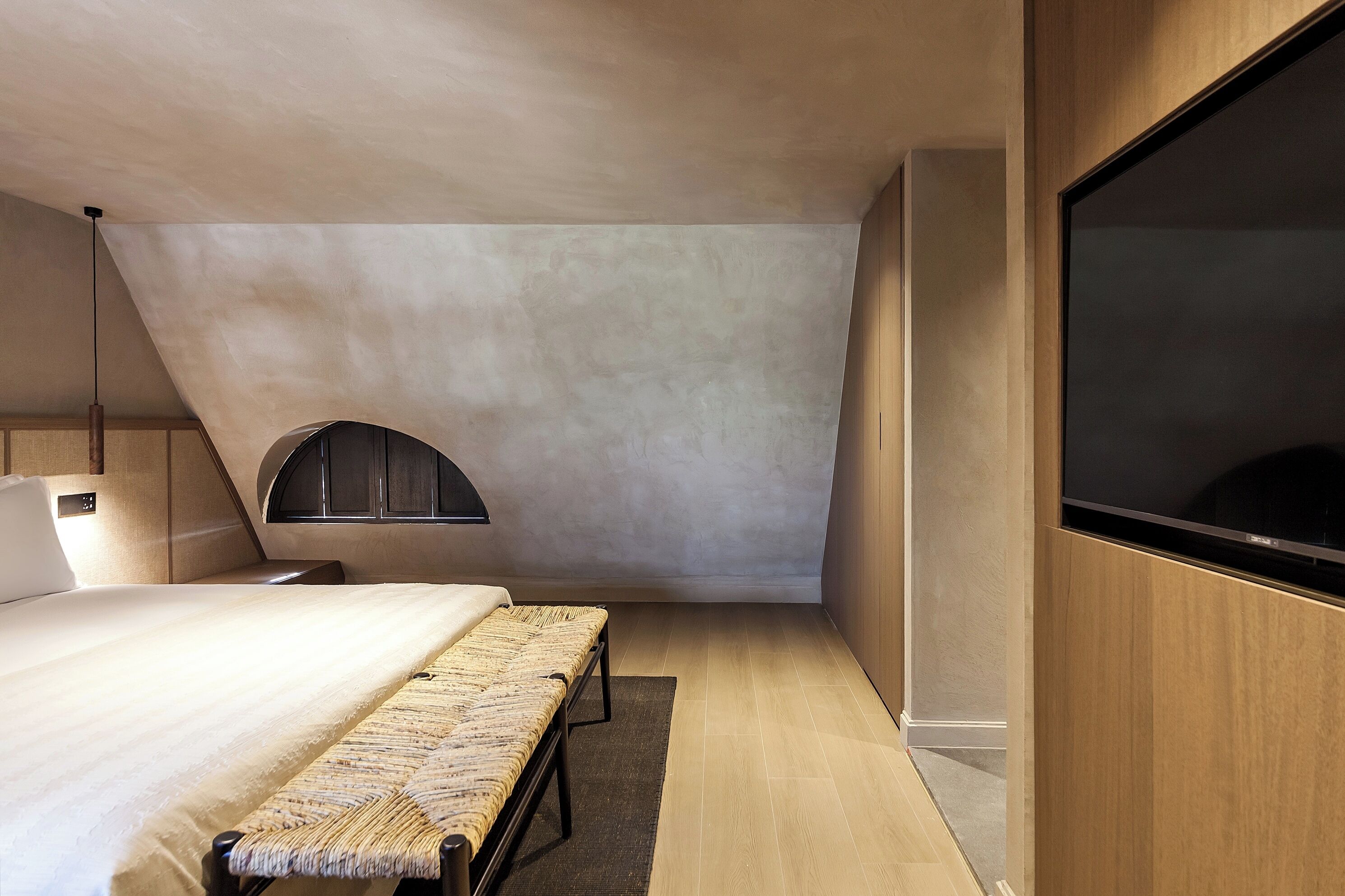 A modern bedroom with a sloped ceiling, a large bed, a bench, a TV, and a small window.
