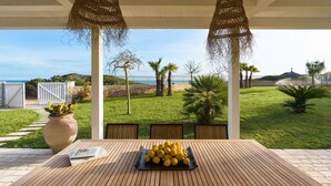 Un tavolo in legno all'aperto con un vassoio di frutta, circondato da sedie, sotto una pergola con cesti fioriti, con vista su un prato curato e un panorama lontano del mare.