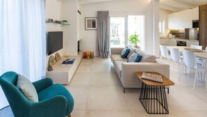 Un soggiorno moderno con un divano, una poltrona color petrolio, un tavolino da caffè e una TV. È presente anche una zona cucina con mobili bianchi e un tavolo da pranzo con sedie.