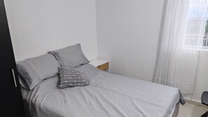 Una cama individual con ropa de cama gris, un almohadón gris con lunares blancos, una mesita de noche de madera y una cortina blanca.