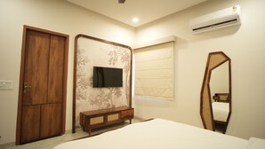 A bedroom with a bed, a wooden wardrobe, a TV mounted on a floral wallpaper wall, a window with blinds, a mirror, and an air conditioning unit. - Angelo's farm (Amer)