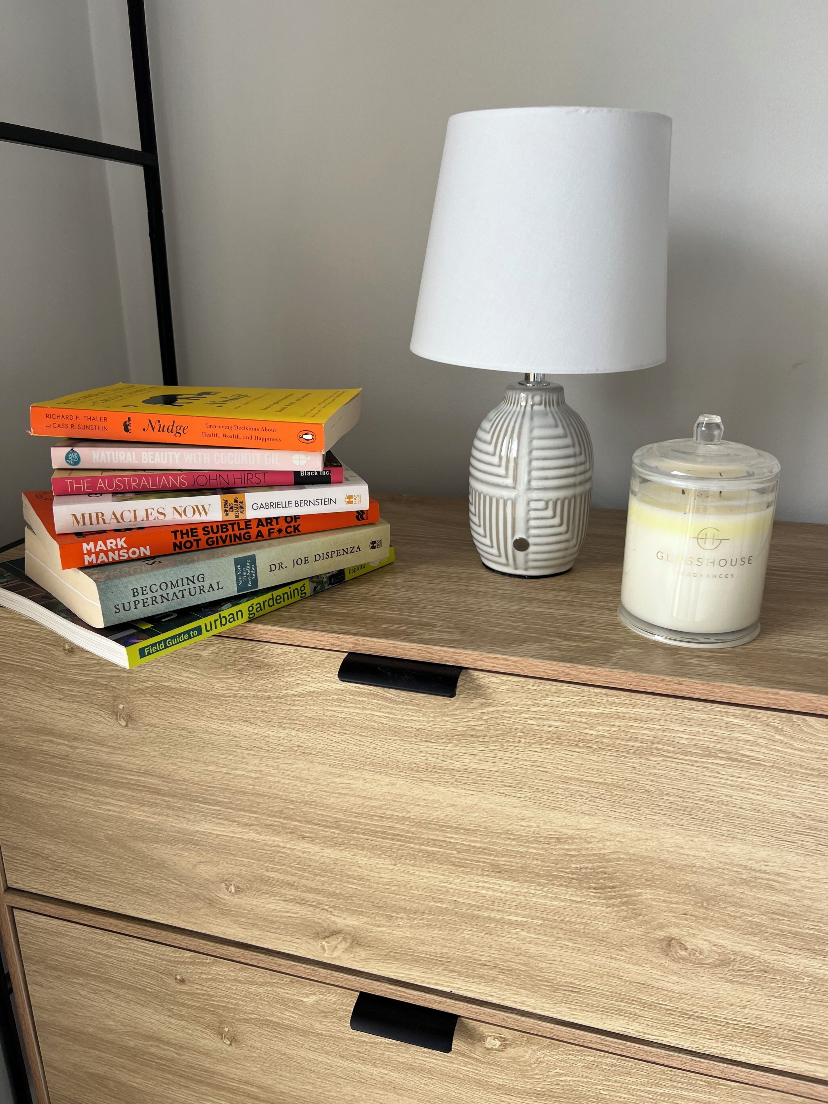 A wooden bedside table with a stack of books, a white lamp, and a glass jar candle.