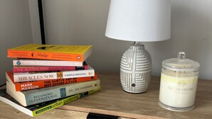 Une table de chevet en bois sur laquelle se trouve une pile de livres, une lampe blanche et un bougeoir en verre.