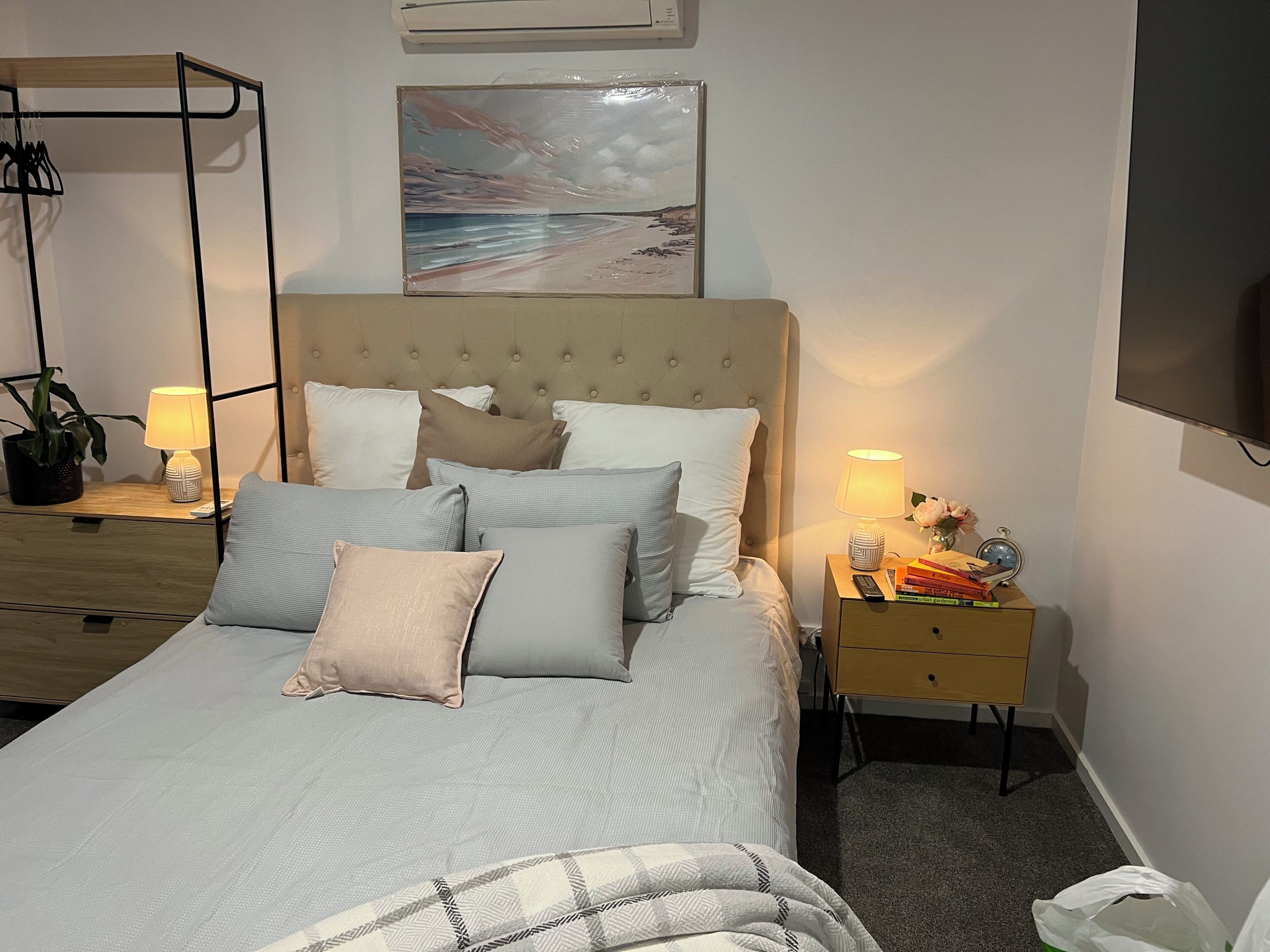 A neatly made bed with multiple pillows, a bedside table with a lamp, a small plant, and a wall-mounted TV.
