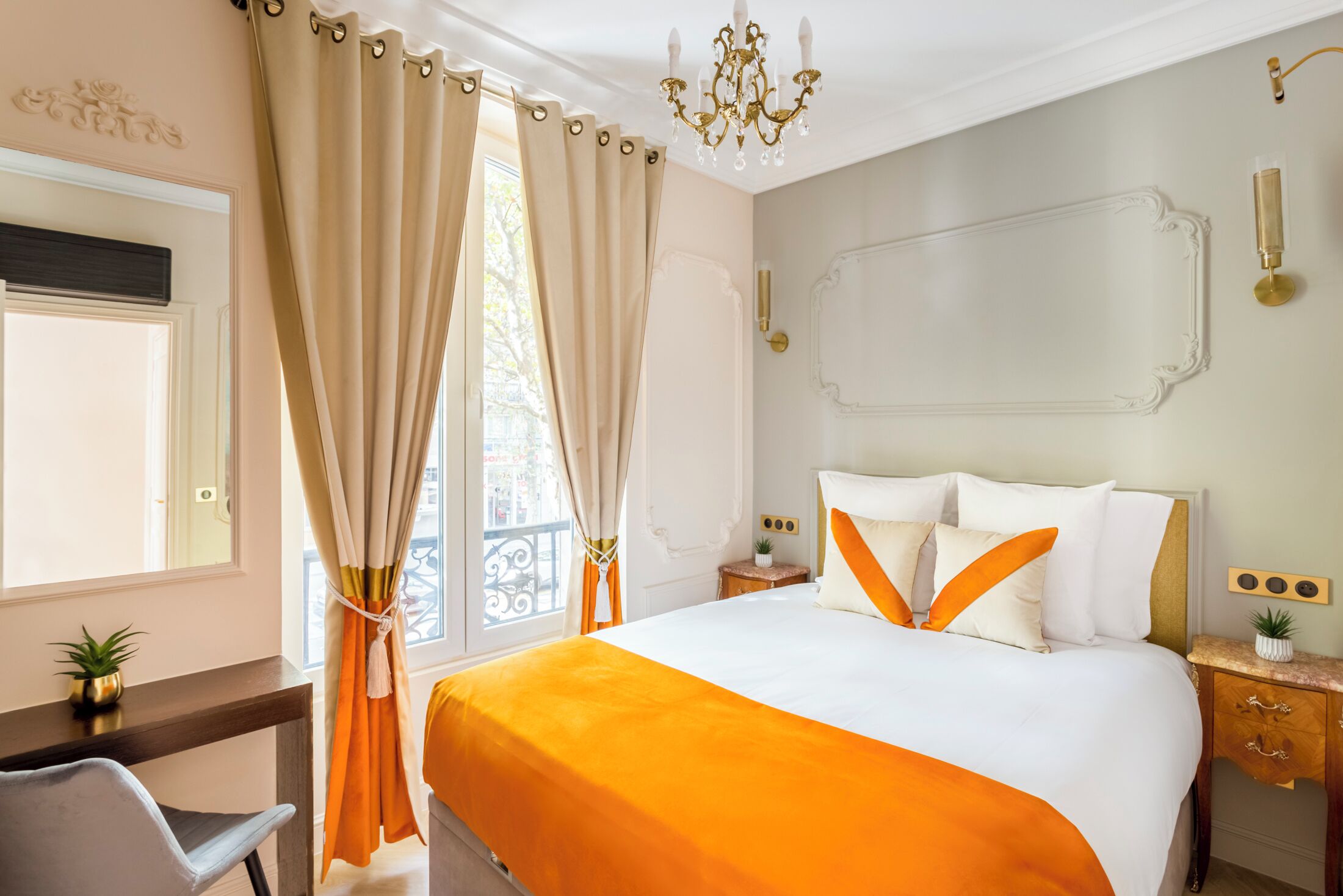 Hotelkamer met een groot bed, een bureau en een stoel. Er is een raam met gordijnen, een kroonluchter en een kleine plant op het bureau.