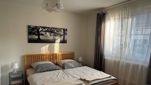 Una cama bien hecha con cabecera de madera, una mesita de noche con lámpara, y un cuadro de árboles en la pared.