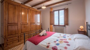 A bedroom with a large wooden wardrobe, a bed with a floral bedspread, a red throw blanket, and a window with a view.