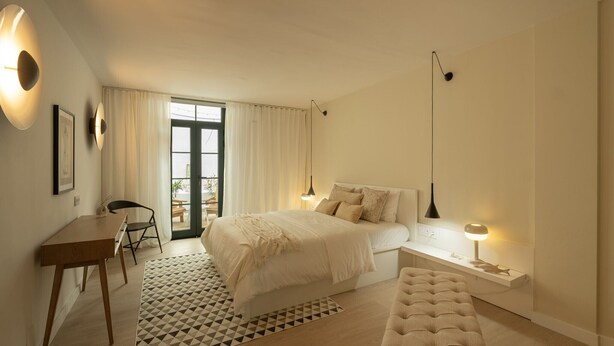A bedroom with a bed, bedside table, lamp, and a view of the outdoors through a window.