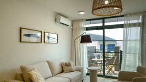 A modern living room with a sofa, a round coffee table, and a chair. There are two framed pictures on the wall, a hanging lamp, and a large window with a view of the outside.