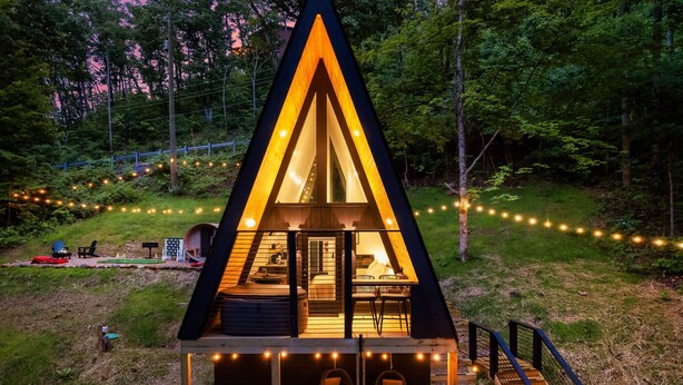 A triangular A-frame cabin with a glass door, a deck with hanging chairs, and string lights. - Treehouse A-Frame, Sauna, Mini Golf, Fully Stocked (Sevierville)