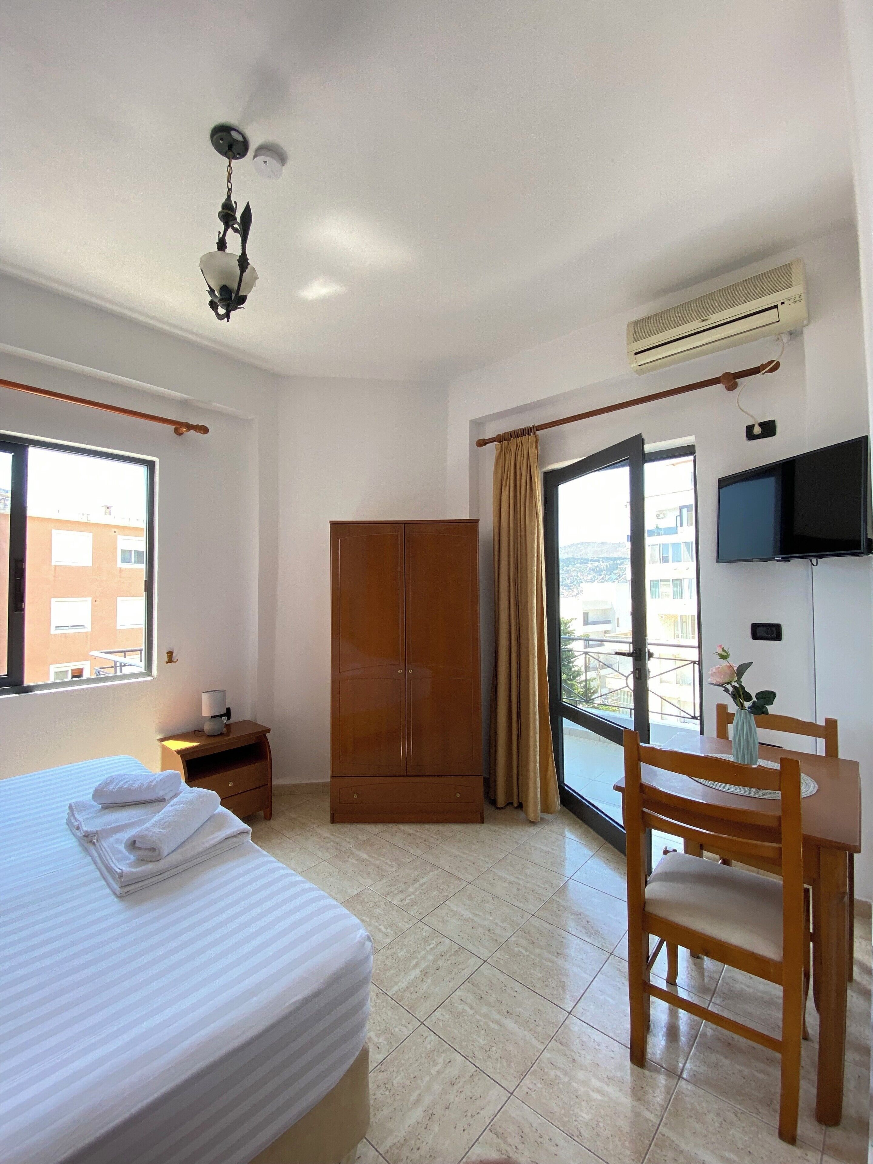 Habitación de hotel con cama, armario de madera, escritorio con silla, televisor y ventana con vista a edificios.