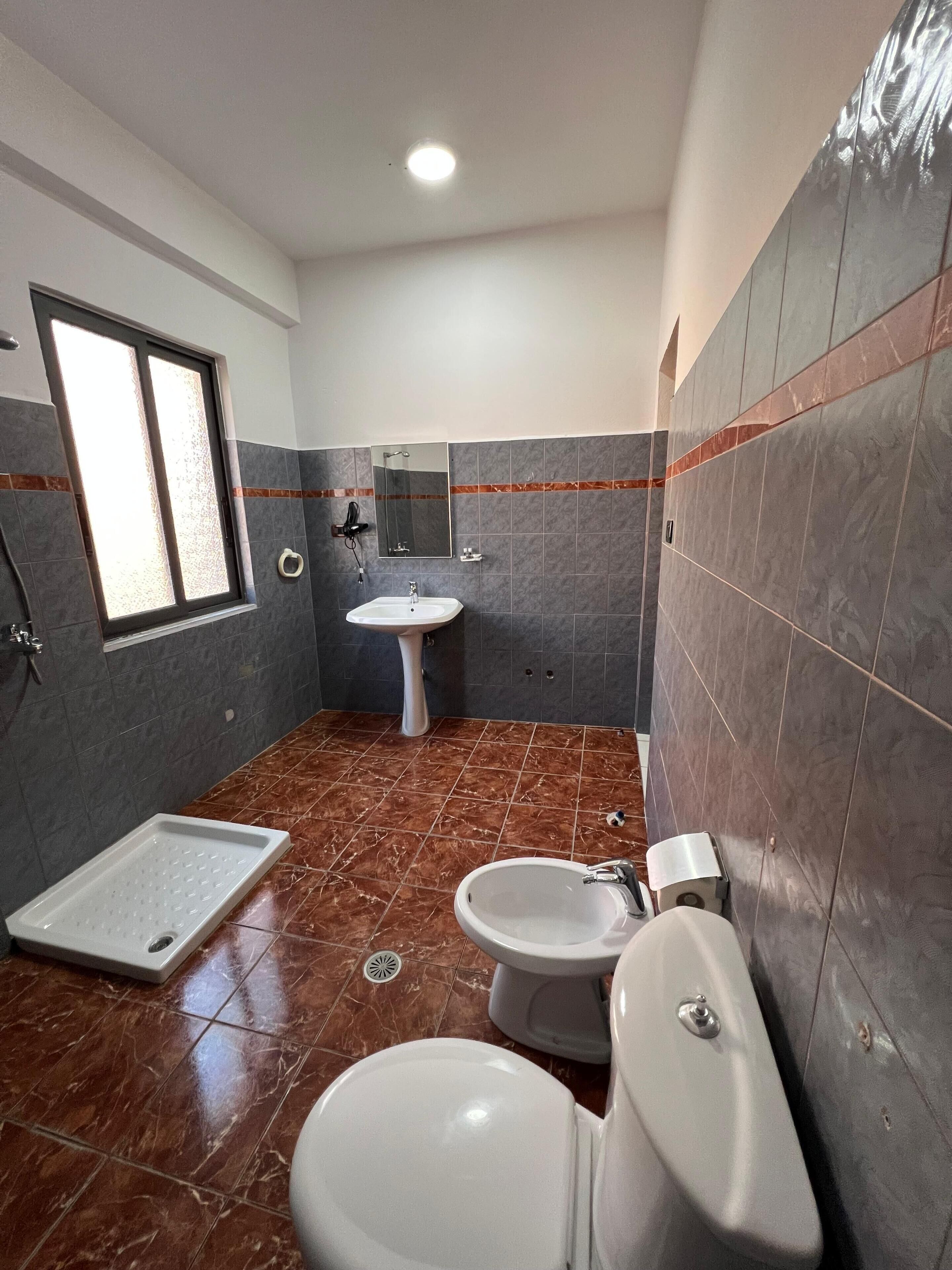 Un baño con inodoro, bidé y lavamanos, paredes de azulejos y una ventana.